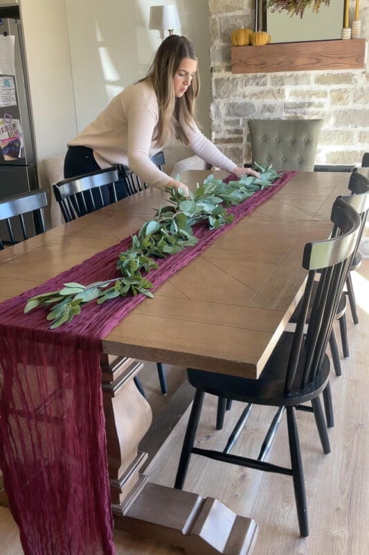 Laying faux eucalyptus garland on a burgundy table runner down a long dining table